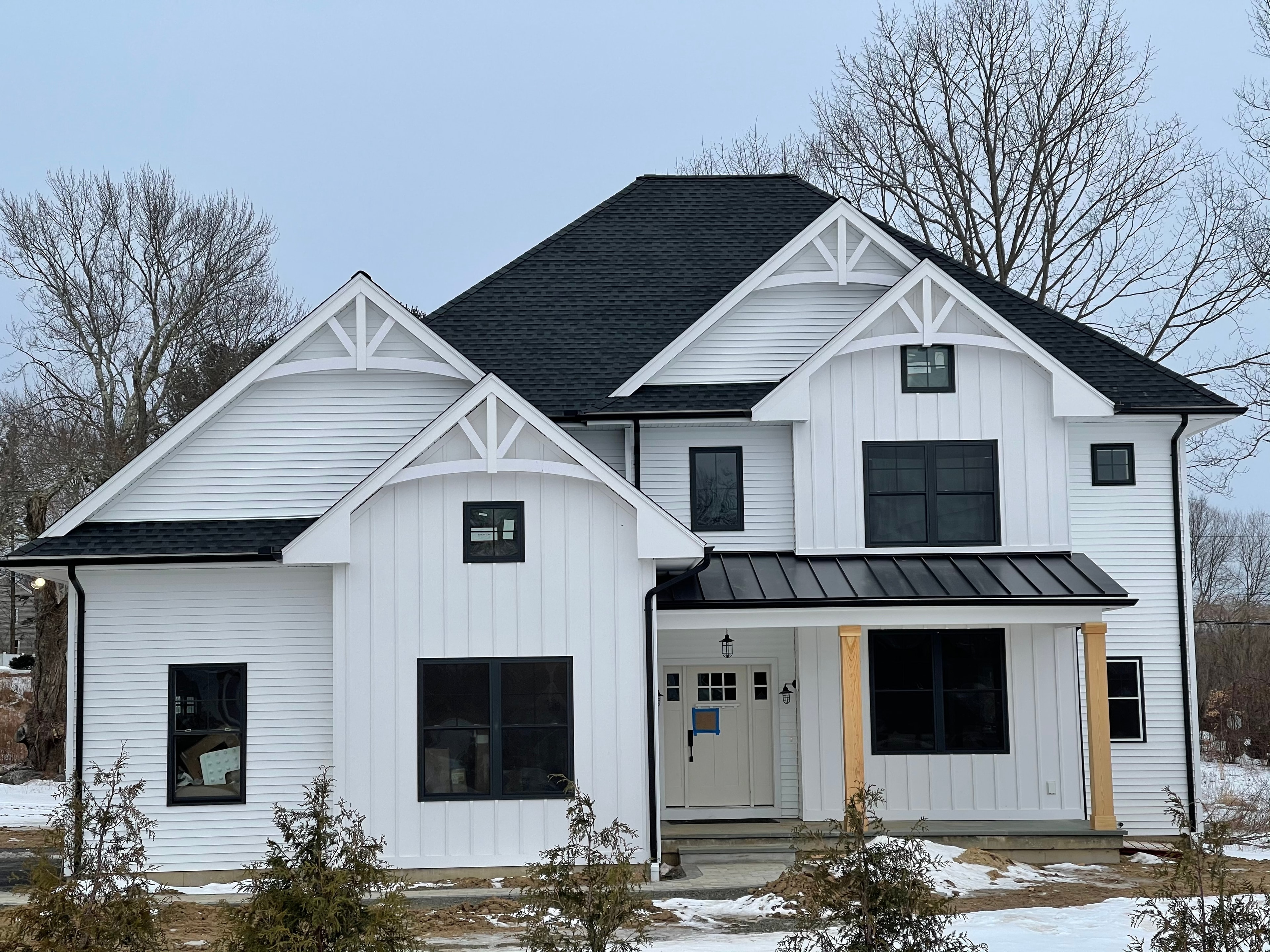 Two-story white farmhouse-style home in vertical board-and-batten siding, with decorative timber brackets in the gables, black-framed windows and a black standing-seam porch roof, photographed from the front on a winter day.